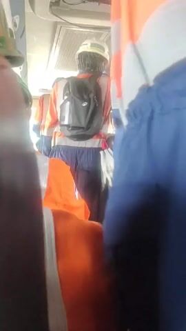 The video shows a view from inside a vehicle, possibly a bus, with several people wearing safety vests and hard hats. The focus is on the back of a person wearing a blue uniform with an orange and white safety vest. A black backpack is visible on their back. Another person in an orange safety vest is partially visible in the foreground. The interior of the vehicle has seats and what appears to be an air conditioning unit or vent on the ceiling. The lighting suggests it is daytime.