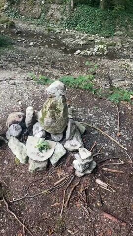 The video captures a nature scene with a dry creek bed in the background, surrounded by trees and greenery. In the foreground, a collection of rocks are arranged in a circular formation, resembling a primitive campfire pit. One large, moss-covered rock stands prominently in the center of the arrangement. Smaller stones are piled around it, and twigs and leaves are scattered on the forest floor. To the left of the rock formation, a black speaker or electronic device is visible. The overall impression is one of a secluded outdoor setting, possibly a place for quiet contemplation or gathering.