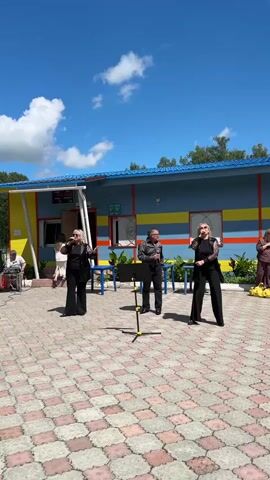 Three people are performing outdoors in front of a colorful building. The two women on either side are dressed in black, with one wearing a lacy overlay on her top. Both women are holding microphones and appear to be singing. The man in the center is also dressed in dark clothing, with a collared shirt, and is holding a microphone. They are standing on a paved area with a patterned surface. A music stand is positioned in front of the man.

The building behind them is painted blue with yellow and red stripes. It has windows and doors, and a sign with some text, some of which is visible. The weather appears to be sunny with a blue sky and scattered white clouds. The time of day is likely daytime. The scene suggests an outdoor performance or event.