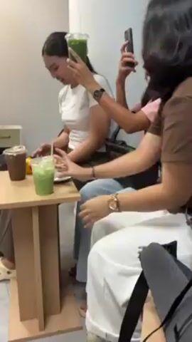 Four women are seated at a table in an indoor space. The table is light brown and holds drinks and a plate of food. One woman wears a white t-shirt and has her hair pulled back. She looks down at the table. Another woman, wearing a pink top and jeans, holds her phone up, seemingly taking a photo or video of the drink. The third woman, in a brown top, is partially visible on the right side of the frame. The fourth woman sits on the right in a white skirt. The environment is plain, with a light-colored wall behind the women and a Marshall speaker visible in the background.