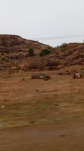 The video shows a landscape of a rocky hill with sparse vegetation. The ground is dry and reddish-brown. Power lines are visible running along the top of the hill. The video appears to have been taken from a moving vehicle. The sky is overcast and grey.