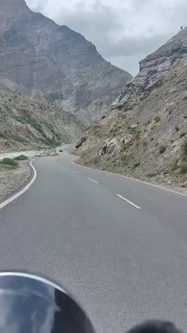A motorcycle is traveling down a paved mountain road. The road is empty except for a few other vehicles visible in the distance. The road curves to the right and is bordered by steep, rocky mountainsides. The sky is cloudy. The camera is mounted on the motorcycle, facing forward.