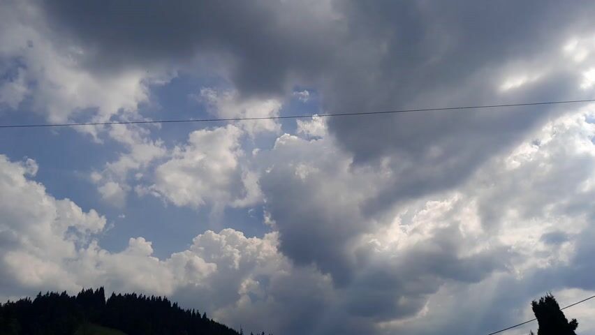 The media file shows a cloudy sky over a forested hillside, likely in Gliczarów Górny, Poland. The scene is set during the day, with sunlight breaking through gaps in the dark, dramatic clouds. The clouds suggest imminent or recent rainfall, creating a dynamic and somewhat somber atmosphere.

The foreground is dominated by a dark silhouette of evergreen trees lining the crest of a hill on the left side of the frame. A single, lighter-colored tree stands on the right, adding a contrasting element. In the upper portion of the image, a thin wire stretches horizontally across the sky, suggesting the presence of human infrastructure, possibly power lines.

The activity is static; it's a snapshot of a natural landscape under a powerful sky. There are no people or animals visible. The focus is on the interplay of light and shadow on the clouds and the dark mass of the forest. The bright patches in the sky hint at a break in the clouds, perhaps a moment of clearing or the sun trying to pierce through. The overall impression is one of a wild, natural landscape under a changeable, moody sky. There is no visible text in the media file.