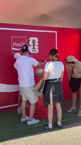 The video shows a group of people gathered around a red display with a Coca-Cola logo and "FIFA WORLD CUP 26" branding. A football is displayed in a glass case on a red pedestal. One person, wearing a white t-shirt and a black baseball cap, is reaching towards the glass case. Another person, in a white t-shirt and a white baseball cap, is standing next to them, also looking at the case. A third person, wearing a beige polo shirt and a beige baseball cap, is looking on from the right side of the frame. A woman in a white t-shirt and a black jacket tied around her waist is standing to the left of the man in the black cap, holding a brown paper bag with a VISA logo. The ground is covered in green artificial turf. The overall scene appears to be an outdoor event or promotion.
