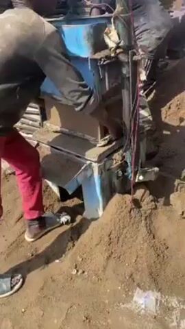 The video shows a group of men using a machine to make cement blocks in an outdoor setting. One man, wearing red pants and a black jacket, removes a fresh block from the machine. Another man, wearing a green tank top, is seen walking near stacks of finished blocks. The environment appears to be a work site with piles of sand, raw materials, and finished blocks. The buildings and trees visible in the background suggest it's a rural or semi-rural area.