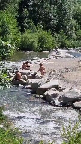 This video captures a sunny day at a riverbank in Setcases, Spain. The scene is dominated by a clear, rocky river flowing through a lush, green landscape. The river has a moderate current, with white water visible in places. Large, smooth stones line the riverbanks, with a sandy area between the water and a concrete retaining wall.

In the foreground, the river flows past a cluster of large, grey rocks. Green foliage is prominent in the lower part of the frame, suggesting a vibrant natural environment.

Further into the scene, a concrete wall runs along the top of the bank, separating the river area from what appears to be a pathway or road. On this upper level, several people are visible. One person is lying on the sandy bank, appearing to sunbathe. Another person is seated nearby, and a third person is partially visible behind them. The presence of swimwear suggests a recreational activity, likely swimming or relaxing by the river on a warm day.

The overall atmosphere is one of a peaceful summer day, with people enjoying the natural beauty and recreational opportunities of the river. The sun is bright, casting shadows, indicating it is likely midday. There is no visible text in the video.