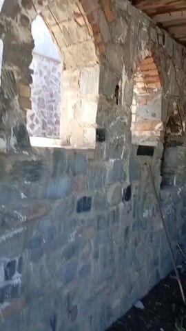 The camera pans around the interior of a stone building with arched windows. The walls are constructed from stacked, uneven stones of various sizes and shades of gray, with some darker stones interspersed. Mortar is visible between the stones. Above each window, there are arched brick details. The roof consists of wooden beams and planks. Debris and tools are scattered on the floor. Through the windows, green foliage can be seen.