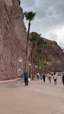 A person is walking down a paved walkway beside a rocky cliff face. There are palm trees planted along the walkway. Several other people are walking in the same direction further down the path. In the background, there are buildings and mountains. The sky is cloudy.