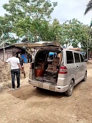 The video shows the back of a silver minivan with its rear door open, parked on a dirt road. A man in a white t-shirt and jeans walks towards the open back of the van and then away again. A second man, also in a white t-shirt and jeans, stands near the open van door. Inside the van, one can see boxes and what appears to be a folded metal structure. A small blue and dark-colored tent is visible on the left side of the frame, next to a makeshift structure. Trees are visible in the background under a cloudy sky.