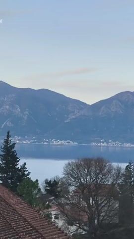 The video shows a view of a bay surrounded by mountains. There are some buildings with red tile roofs in the foreground. The water is calm, and the sky is a pale blue with some light clouds. The mountains are dark and covered with trees. There are some small towns visible on the far side of the bay.