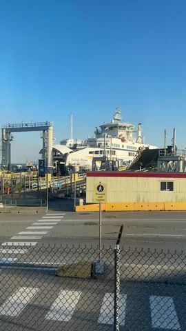 The video shows a ferry docked at a terminal. The ferry is named "Salish Heron". There are ramps leading from the terminal onto the ferry, indicating that vehicles can drive on and off. The area around the ferry terminal appears to be an outdoor port with a clear blue sky. There is a pedestrian crossing marked on the road leading to the ferry. A sign with a pedestrian symbol and the words "Proceed With Caution" is visible. A chain-link fence is in the foreground, partially obscuring the view.