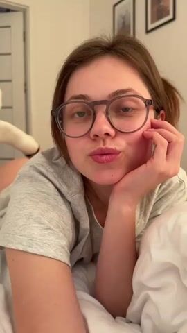 A young woman with brown hair and glasses lies on a bed, propped up on her elbow. Her hand is supporting her chin. She is wearing a grey t-shirt and white socks. The room is dimly lit and appears to be a bedroom. There are framed pictures on the wall behind her. The woman makes various facial expressions, including looking off to the side and smiling at the camera.