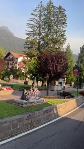 The video shows a roadside view of a village, likely in a mountainous region. The scene is captured from a moving vehicle, suggesting a perspective from a car or bus. On the left side of the frame, there is a mature tree with dark red foliage, a small wooden kiosk, and park benches. Beyond the trees, buildings with pitched roofs are visible. To the right, a lamppost with a sign displays a map with various directional arrows. Further along, a crosswalk is marked on the asphalt road. There are flower beds with red and white flowers along the roadside. The background shows more buildings and lush green trees, with the silhouette of a mountain peak in the distance. The sky is clear and bright, indicating it is daytime. No people are visible in the video.