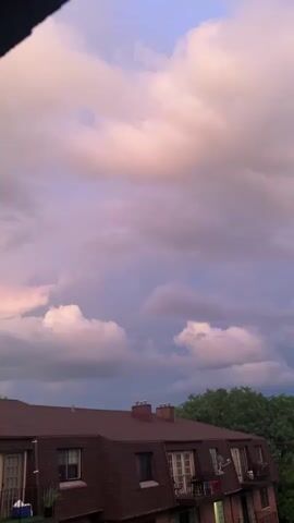 The video shows a dusky sky with dramatic clouds above a building. The clouds are illuminated by the setting sun, displaying hues of pink, purple, and gray. The building below has a dark brown, mansard roof with multiple dormer windows and balconies. Trees are visible to the right of the building. The video is static and appears to be filmed from a high vantage point, possibly a balcony or window. There are no people or actions visible in the frame.