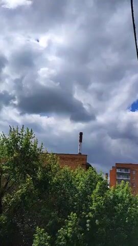 A vertical video captures an outdoor scene with a cloudy sky. In the foreground, lush green trees obscure the lower part of the frame. Above the trees, a brick building is visible with a tall, red and white striped chimney extending upwards. Further in the background, another building with multiple windows can be seen. The clouds are predominantly gray, suggesting overcast conditions, but patches of blue sky are also visible. The camera appears to be stationary.