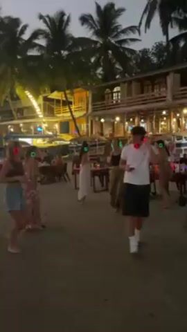 The scene takes place on a beach in the evening, with palm trees silhouetted against the sky. Several people are dancing on the sand. They are wearing wireless headphones that light up in different colors (red, green). Some people are seated at tables, some of which are lit by candles. A building stands in the background. A man walks past, looking at his phone.