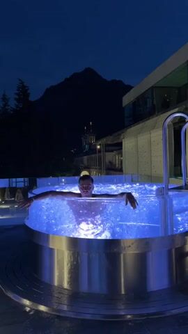 A person is submerged in a transparent hot tub illuminated by blue lights. The person is looking at the camera with their hands pressed against the glass. The hot tub is outdoors at night, with a dark mountain and a building in the background.
