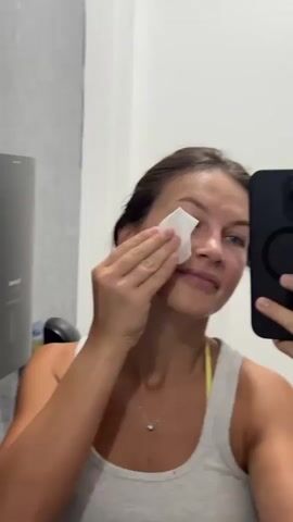 A woman with brown hair, wearing a grey tank top and a yellow sports bra, is seen in a bathroom, holding a black smartphone in her right hand and a white wipe in her left hand. She is using the wipe to clean her face, specifically her cheek and forehead. A mirror is visible in the background, reflecting her image and the surrounding bathroom environment, which includes a grey tiled wall and a black dispenser. The woman is looking at the camera, which is being held by her phone.