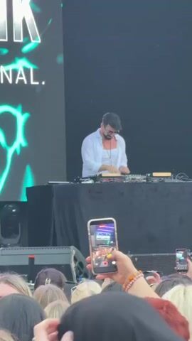 This video shows a DJ performing on a stage with a large screen behind him. The DJ is a man wearing a white shirt and sunglasses. He is standing behind a DJ booth with turntables and other equipment. In front of the stage, a crowd of people are watching the performance. Some people in the crowd are holding up their phones to record the DJ. The screen behind the DJ displays text that reads "KAN IŞIK" and "L. INTERNATIONAL." The overall atmosphere appears to be that of an outdoor music festival or concert.