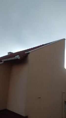 The video shows the exterior of a house on a cloudy day. The house has a beige stucco exterior and a red tiled roof. A small white window is visible on the side of the house. There is also a clothesline attached to the side of the house. In the background, there are trees and a dark sky. The overall mood is somewhat somber due to the overcast weather. The time of day appears to be late afternoon or early evening, given the subdued light. There is no visible text in the video. The location context is Zrenjanin, Serbia. The activity taking place is simply the passive observation of the house and its surroundings.