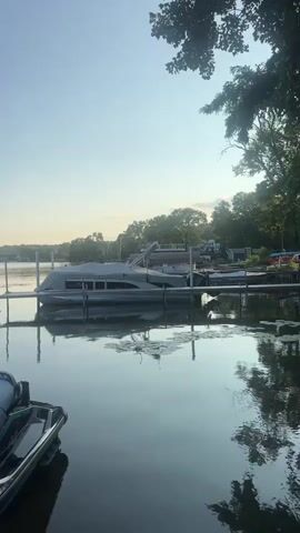 The video begins by showing a lake with boats docked. The camera then pans to a jet ski in the foreground. The camera continues to pan to the right, showing more boats docked and a sign that says "Happiness is a Boat". The video ends showing the lake with boats docked and trees in the background. The sky is clear and blue.