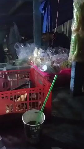 The video shows a poorly lit outdoor food stall. A person is visible in the background, tending to the stall. There are several red and blue plastic baskets, a large metal kettle, and various containers. A cup with a green straw sits in the foreground. The sound of a wailing siren is present throughout the video.