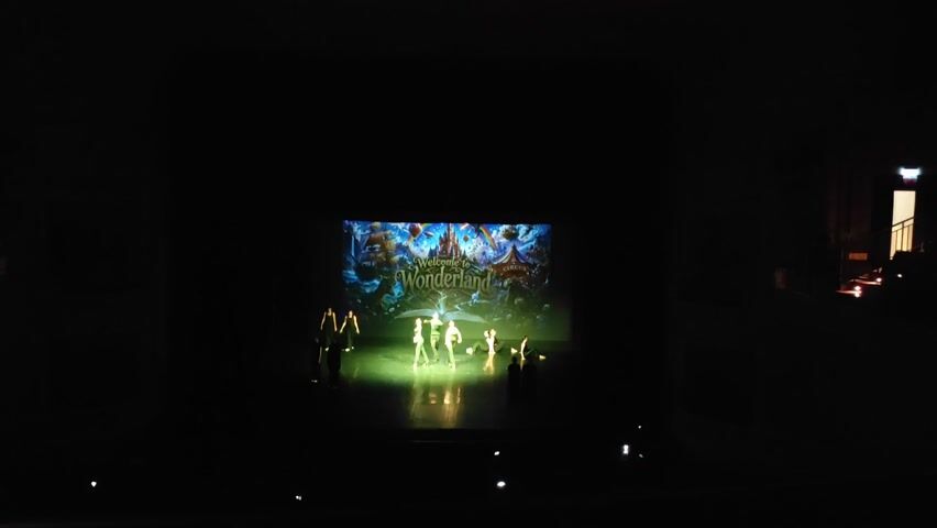 The video shows a stage performance in a dark theater. A projected image of a castle with the words "Welcome to Wonderland" is displayed on a screen at the back of the stage. A group of dancers, dressed in light-colored costumes, are performing in front of the screen under a spotlight. The dancers are moving in unison and at times are in various formations. The stage floor reflects the light from the performance. In the foreground and scattered around the lower part of the frame, there are small, blurry lights, possibly from other audience members' phones or electronic devices. To the right of the stage, there is a glimpse of a dimly lit hallway or backstage area with some seating visible.