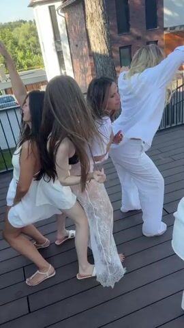 Four women are dancing on a wooden deck, likely a balcony or rooftop. They are all wearing white outfits. From left to right, the first woman has long dark hair and is wearing a white sundress. The second woman has long brown hair and is wearing a black top and a white skirt. The third woman has brown hair and is wearing a white t-shirt and a sheer white lace skirt. The fourth woman has blonde hair and is wearing a white shirt and white pants.

The scene appears to be a daytime event, possibly a party or celebration, given the festive atmosphere and dancing. The sun is out, suggesting pleasant weather. In the background, there is a modern house with a brick facade and a pitched roof, surrounded by lush green trees and vegetation. A railing runs along the edge of the deck. A man in a white t-shirt and shorts is visible in the background on the right, also appearing to be at the event. The overall mood is joyful and energetic, with the women fully immersed in their dancing.

No visible text is present in the media. The context provided indicates the location is Cojușna, Moldova.