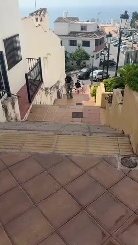 A vertical, eye-level, medium shot shows a set of outdoor stairs leading down toward a town and the sea. The stairs are made of light brown tiles and yellow textured tactile paving. Two people are descending the stairs. The person in the foreground is wearing a camouflage dress and dark sandals, and is walking away from the camera. The second person is farther down the stairs and is also walking away from the camera. To the left of the stairs is a white wall with a black metal railing. To the right of the stairs is a yellow planter box with green plants. In the background, there are white buildings, cars parked along a street, street lamps, and the sea under a cloudy sky.