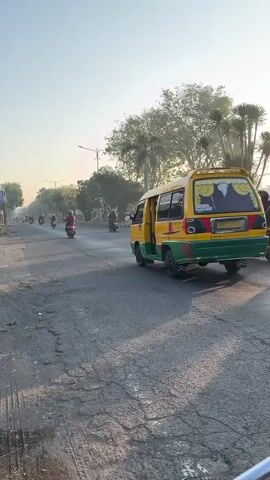 The video shows a busy street scene during daylight hours. Several motorcycles are traveling in the same direction, with one in the foreground and others further down the road. A yellow and green van is also visible, moving along the street, and appears to be emitting smoke from its exhaust. There are trees lining the side of the road, and the sky is clear with a warm, hazy glow, suggesting it might be early morning or late afternoon. The road surface appears to be asphalt and shows signs of wear and tear, with some cracks and discoloration. The overall atmosphere suggests a typical urban environment with active traffic.