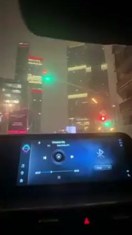 The video shows a car driving through a city at night during rain. The dashboard with a touchscreen display is in focus, playing a song. Skyscrapers with illuminated windows and lights are visible through the windshield, and traffic lights are lit. The driver's hands are visible on the steering wheel, which features digital gauges. The overall scene suggests an urban night drive in wet weather.