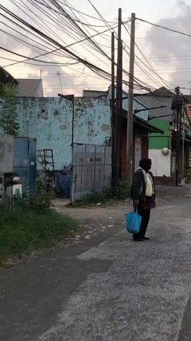 The video begins with a wide shot of a street scene, with a person standing on the right side of the frame, holding a blue bag. The person is wearing a dark jacket over a yellow shirt and a red skirt, and a black hijab. They are facing to the right. The background features a dilapidated blue wall with some green elements and overhead wires. The shot then zooms in on the person, who turns to face the camera. They are wearing glasses, and the yellow shirt appears to be a uniform with a badge or emblem on the left chest. The person is holding the blue bag in their left hand and a red purse is slung across their body. The person appears to be walking slightly towards the camera. The environment looks like a somewhat neglected urban or industrial area, with rough pavement and overgrown vegetation in places. The lighting suggests it might be late afternoon or early evening.
