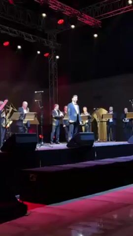 The video shows a man in a suit singing on a stage with a band. The band consists of five men, dressed in suits, playing instruments including saxophones, a flute, and a clarinet. The stage is dark with red spotlights shining down. In the background is a building with the word "Adda" visible on the wall. A truss with more lights is visible above the stage. There are speakers along the front edge of the stage. Someone is throwing a yellow balloon at the beginning of the video.