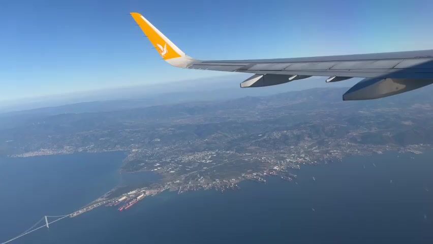 The video shows an aerial view from an airplane window, with the wing of the plane prominently in the foreground. The wing is white with a yellow logo on the wingtip. Below the wing, a vast blue body of water stretches out, meeting a coastline dotted with buildings and a large port. Several ships are visible in the water and docked at the port. A long bridge or causeway extends into the water from the coastline. In the background, rolling hills covered in vegetation and trees rise up, meeting a clear blue sky. The overall scene suggests an airplane flying over a coastal city and its harbor under a bright sky.