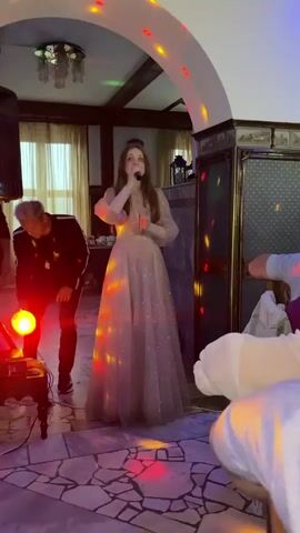 A young woman in a sparkling, pale pink gown with puffed sleeves sings into a microphone on a tiled floor. She is the central subject, illuminated by colorful disco lights projected onto the floor and walls, creating a festive atmosphere. Behind her, a man in a black jacket and pants is bending down near a speaker and a small table with a glowing sphere. In the background, there are draped curtains, wooden beams on the ceiling, and ornate wall decor. To the right, partially visible figures are seated, also enjoying the event. The lighting suggests it is nighttime or an indoor event. The overall scene depicts a performance or celebration, possibly a party or concert, in an indoor venue. There is no visible text. The location context provided is Zavod Mikhailovskii, Russia.