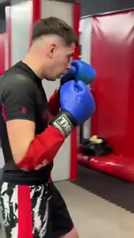 In a gym setting, a man wearing boxing gloves (red on the left hand and blue on the right) is shown boxing. The gym has a red punching bag attached to a red wall. The man is wearing a black and pink top and black and white patterned pants with red detailing. The Belarusian flag is visible on the wall. Another person, wearing a white shirt and head gear, is seen briefly assisting with the training.