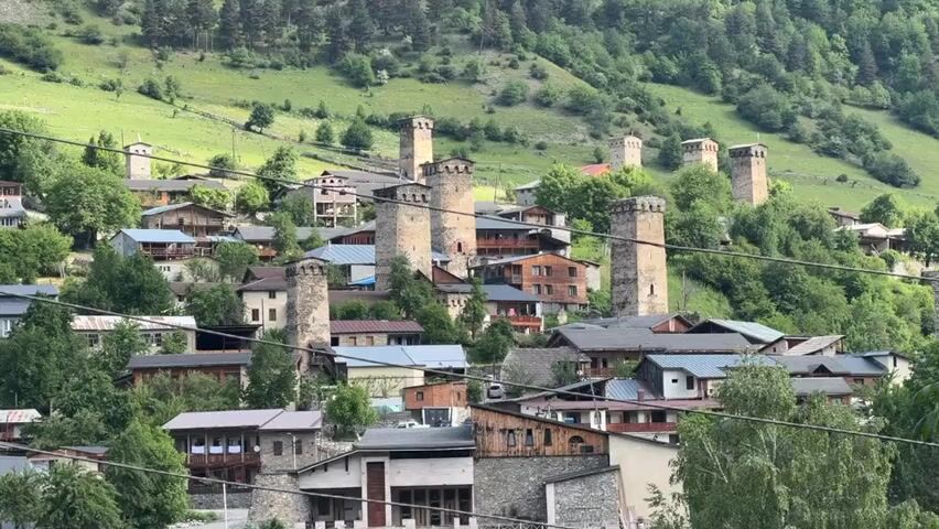 A distant view of a village nestled in a green, hilly landscape. The village is characterized by numerous stone towers and buildings, many of which appear to be traditional Svan towers, some with attached wooden structures. The buildings are clustered together on the hillside, with trees and vegetation interspersed throughout. The sky is overcast. The focus is on the architecture and the natural surroundings.