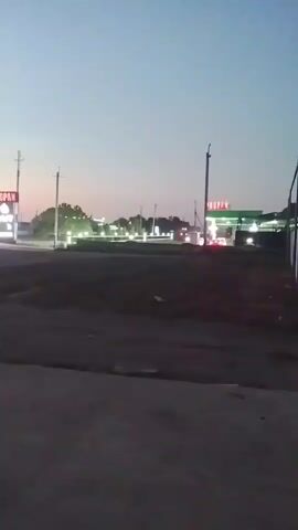 The video shows a gas station at dusk. The sky is a pale blue with hints of pink near the horizon. There are several signs visible, some with illuminated text that is difficult to read. A gas pump canopy with lights is also visible. The area appears to be relatively flat and sparsely vegetated, with some trees in the distance. The ground in the foreground is paved. The lighting suggests that it is either early morning or late evening.