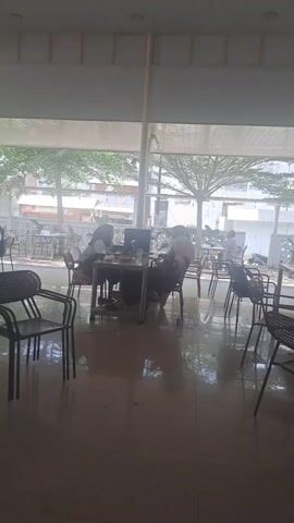 The video shows an indoor/outdoor restaurant setting with tables occupied by people who appear to be eating and drinking. Several tables and chairs are visible in the foreground, some stacked. The area has a polished tile floor that reflects light. Large glass windows offer a view of the outside, where there are trees, parked vehicles, and buildings. The ceiling has recessed lighting and ceiling fans. The setting seems casual and populated with people during the daytime.