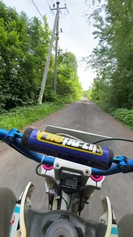A first-person perspective video captures a person riding a dirt bike along a tree-lined dirt path on a sunny day. The camera is mounted on the handlebars, showing the blue handlebars with a black and yellow "PROTAPER" pad, and the white front fender of the bike. The path is narrow and flanked by lush green trees and bushes on both sides. In the distance, the path appears to curve slightly to the right. The sky is clear blue with a few wispy clouds. Power lines and utility poles are visible on the left side of the path at the beginning of the video. The dirt path is uneven and appears to have loose gravel. The bike is moving forward steadily, and no other vehicles or people are visible.