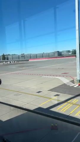 The video shows an airport tarmac viewed through a window. The tarmac is marked with yellow and red lines and cones. In the distance, there are large buildings with red and white stripes. A jet bridge extends from the terminal building, and a small tug vehicle is parked nearby. The sky is clear and blue. The camera pans across the scene, offering different perspectives of the airport environment.