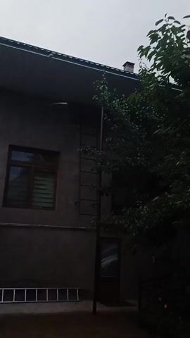The video shows a dark, overcast day in Angren, Uzbekistan. The scene is an exterior view of a building, likely a house, with a dark grey, textured wall. To the left, a window with horizontal blinds is visible. Below the window, a silver ladder is laid out on the ground. A metal ladder runs vertically up the side of the building, attached to the wall. To the right of the building, a large tree with dark green leaves partially obscures a doorway. A chimney can be seen on the roof of the building. The overall impression is one of a quiet, potentially damp day, with an emphasis on the functional aspects of the building and its surroundings. No people are visible, and the focus is on the architecture and natural elements. The lighting suggests it is either early morning or late afternoon, or simply a very cloudy day.