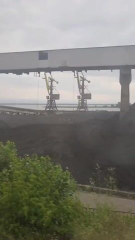The video pans across an industrial area with large piles of dark material, likely coal, on the ground. Two large cranes are visible in the distance, and a covered conveyor belt system stretches across the scene. The area appears to be near a body of water, possibly a river or lake. Vegetation, including trees and grass, is growing around the industrial structures. In the foreground, train tracks are visible. A white car is parked near a concrete structure in the middle ground. The sky is overcast.