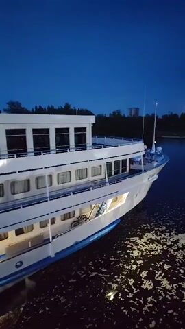 The video shows a white cruise ship sailing on dark water at dusk. The ship has multiple decks with rows of windows. Lights are visible inside the ship, illuminating some of the cabins. The sky is dark blue. Trees and a distant building are visible on the shoreline. Two people are sitting on a deck of the ship.