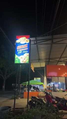 The video captures an outdoor food stall area at night. A brightly lit sign for "SEBLAK & RUMAH MAKAN" (Spicy Noodle Soup & Eatery) is prominently displayed on the left. The area is covered by a metal roof supported by poles, with strings of lights illuminating the space. Several motorbikes are parked in front of the stalls. In the background, various food stalls are visible, some with people working behind the counters. One stall displays food items and has a sign in Indonesian. Another stall, "ES UBI UNGU CREAMY DEK NALA," advertises purple sweet potato ice cream. The ground appears to be paved and somewhat uneven. Trees are visible on the far left, and electrical wires crisscross the sky overhead. The overall atmosphere suggests a bustling night market or food court setting.