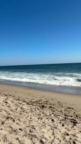 A wide shot of a sandy beach on a clear day. The ocean is calm with gentle waves rolling onto the shore. The sky is a vibrant blue. In the distance, a person is walking along the water's edge. The sand in the foreground is textured with footprints and small indentations.