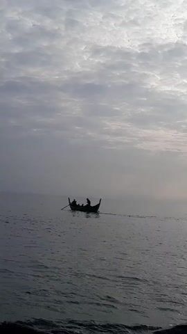 The video depicts a small boat moving on the water. The sky is overcast. In the boat, several figures are visible. One person is standing and reaching toward the center of the boat, while another is seated and operating a long oar. The boat appears to be traveling away from the viewer.