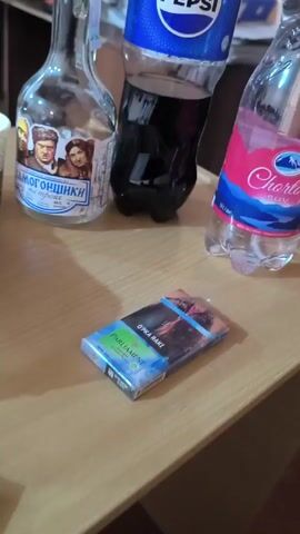 The video shows a table with a pack of Parliament cigarettes, a bottle of vodka, a bottle of Pepsi, and a bottle of Chortoq mineral water. The camera pans to reveal a mostly eaten slice of pizza in a plastic bag, and an empty glass. In the background, a person is seated, using a cell phone.