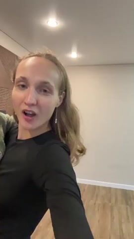 The video opens with a woman holding a baby in her arms in a room. The woman has her mouth open in an excited expression. The baby is wearing a green outfit. The room appears to be a living room with a wooden accent wall, a television, and a white entertainment unit. The woman then begins to spin around, holding the baby securely. Her hair flies around her head as she spins. The baby also spins with her, appearing to be enjoying the ride. The video continues with the woman and baby spinning for a few seconds before the clip ends.