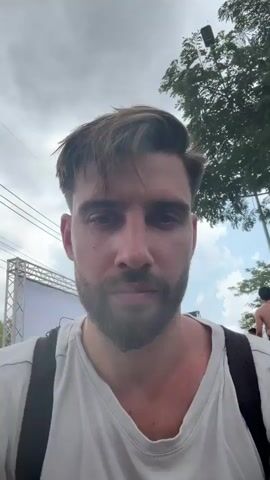 A bearded man with brown hair and a white v-neck shirt is looking down at the camera, possibly recording a selfie video. He has a black backpack on. The background shows what seems to be an outdoor festival or event, with stage structures and other people around, including a shirtless man walking in the distance. Trees and a cloudy sky are also visible.